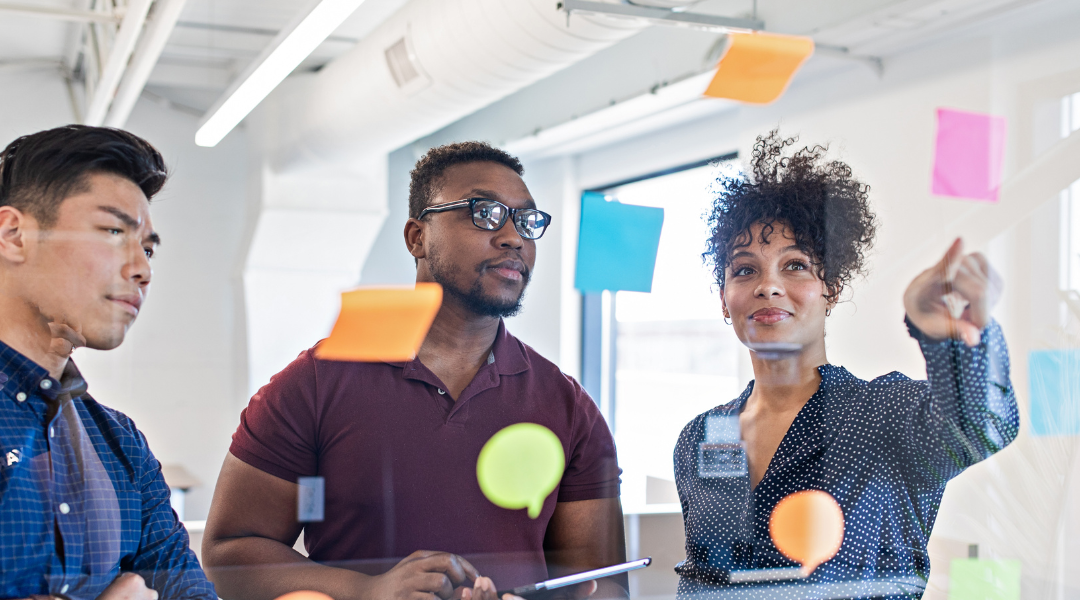 A woman and two men looking at a clear board covered with brightly colored sticky notes.