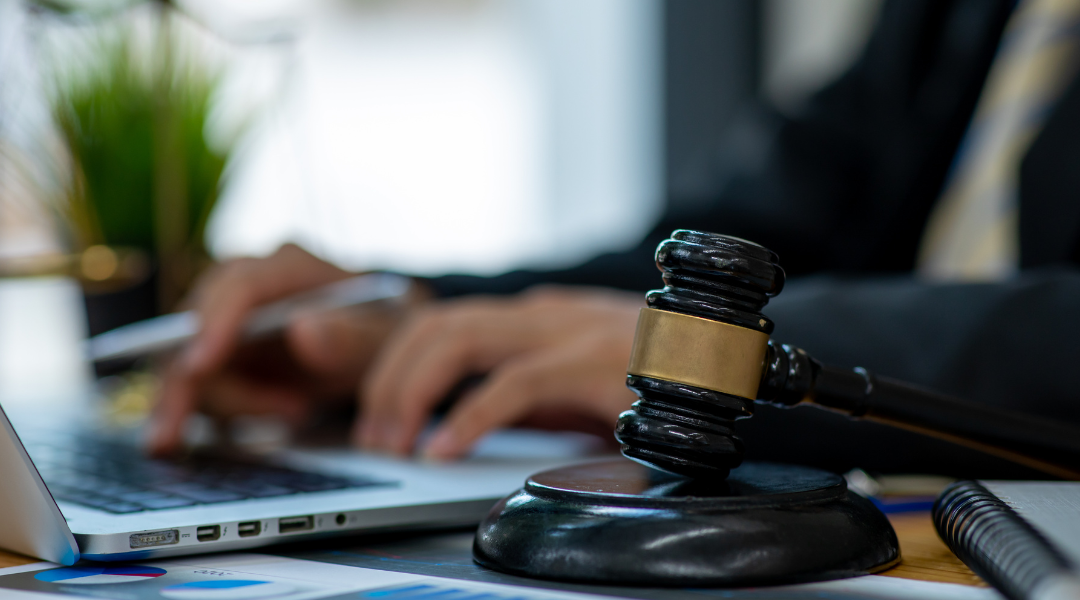 A desk with a laptop and a gavel, while a person in a black suit and yellow-and-blue striped tie types on the laptop.
