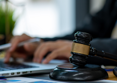 A desk with a laptop and a gavel, while a person in a black suit and yellow-and-blue striped tie types on the laptop.