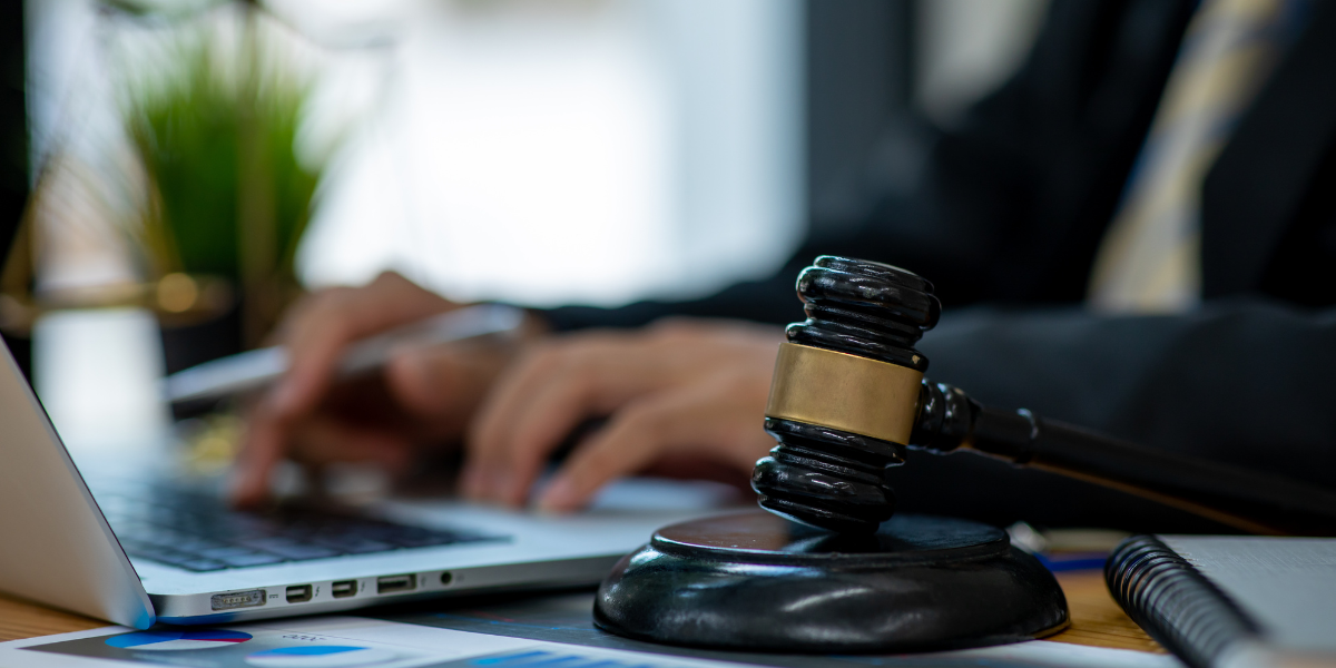 A desk with a laptop and a gavel, while a person in a black suit and yellow-and-blue striped tie types on the laptop.