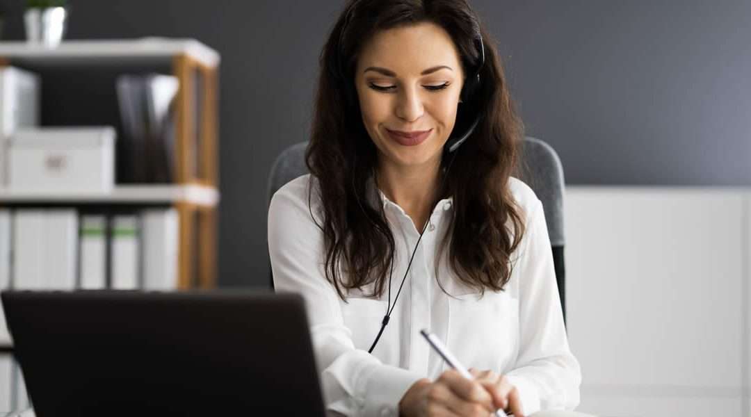 A woman in a black office wearing a headset and working at a laptop.