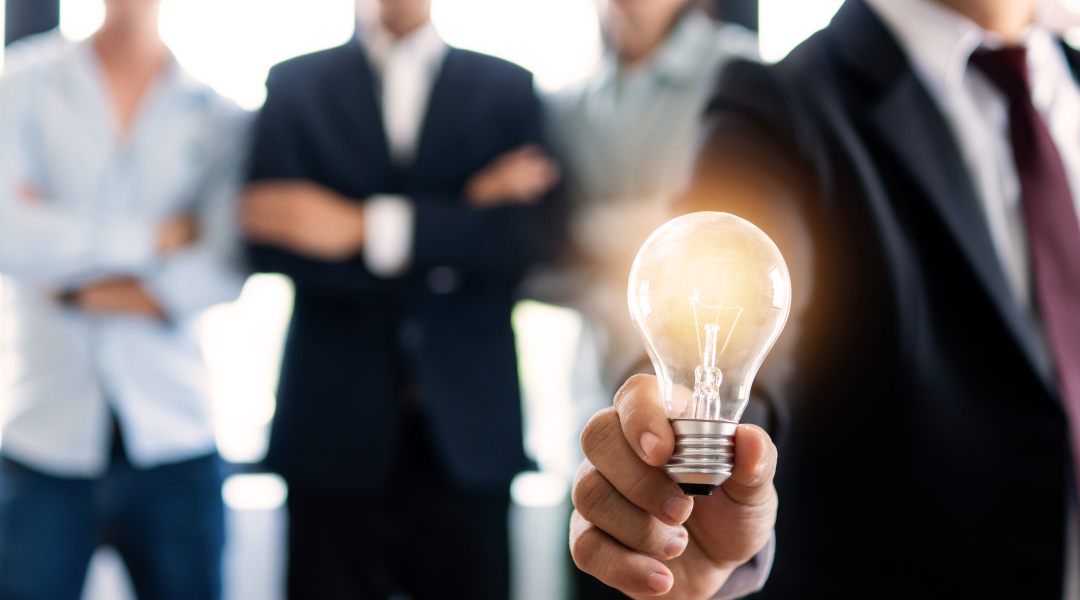 A man in a black business suit and maroon tie holding a lightbulb, with three men lined up in the background.