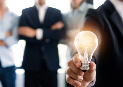 A man in a black business suit and maroon tie holding a lightbulb, with three men lined up in the background.