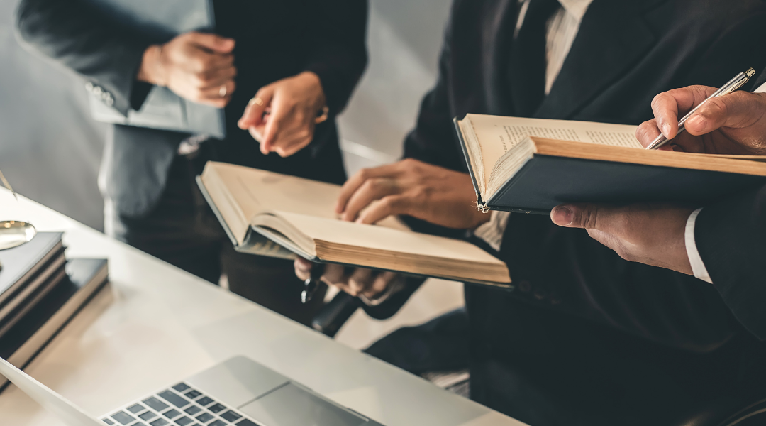 Close-up of three men in black suits holding legal books, with a laptop and scales of justice on the table in front of them.