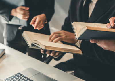 Close-up of three men in black suits holding legal books, with a laptop and scales of justice on the table in front of them.