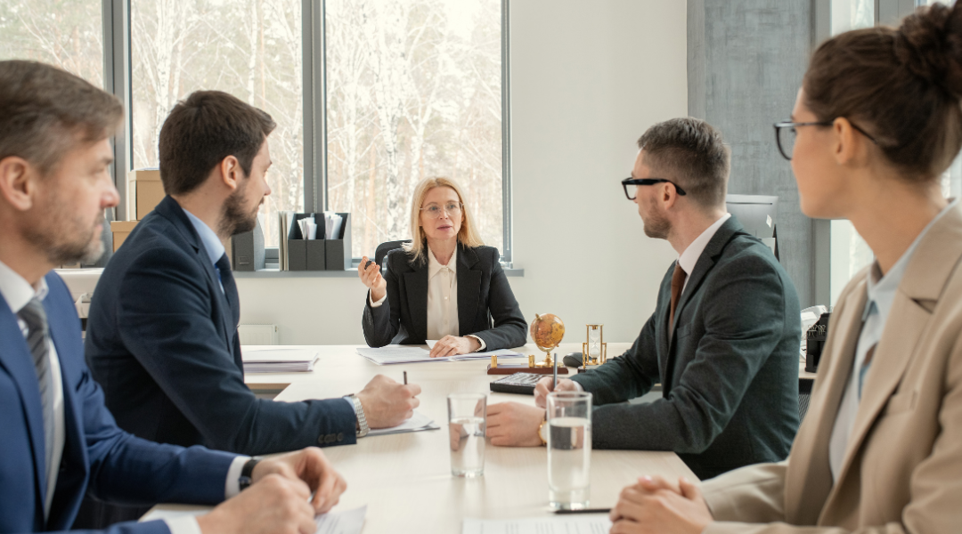 Three men and two women in business suits, talking seriously at a conference table.