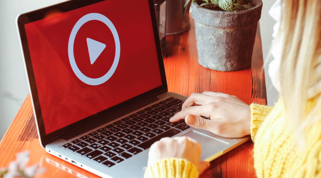 A woman with blonde hair, wearing a yellow sweater, at a laptop with a red screen and a large white play button.