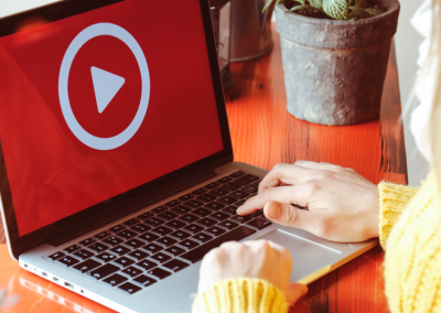 A woman with blonde hair, wearing a yellow sweater, at a laptop with a red screen and a large white play button.