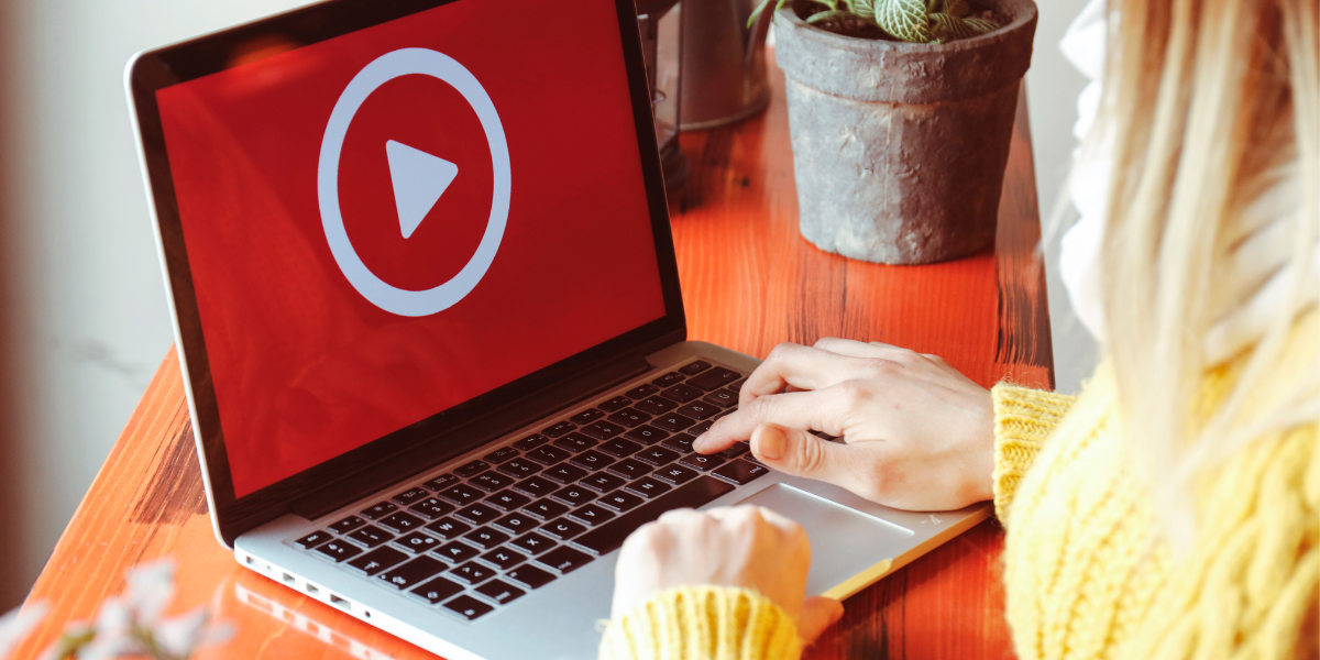 A woman with blonde hair, wearing a yellow sweater, at a laptop with a red screen and a large white play button.