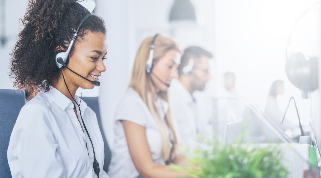 Two women and one man wearing headsets in a brightly lit call center office, with a green plant in the foreground.