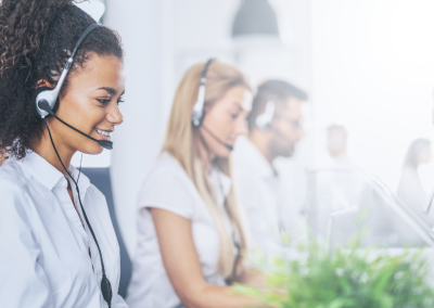 Two women and one man wearing headsets in a brightly lit call center office, with a green plant in the foreground.