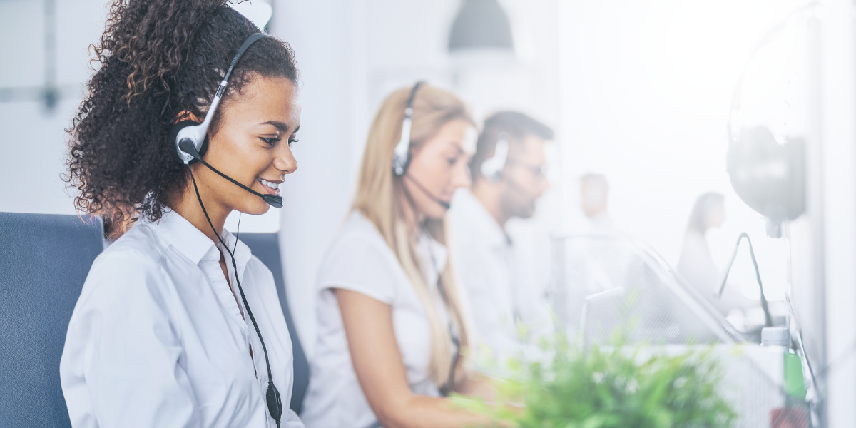 Two women and one man wearing headsets in a brightly lit call center office, with a green plant in the foreground.