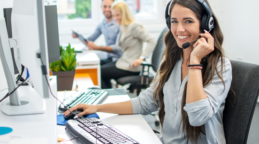 A smiling woman wearing a headset, sitting at a computer in a brightly lit office.