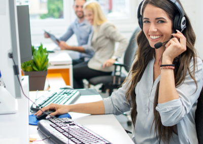 A smiling woman wearing a headset, sitting at a computer in a brightly lit office.