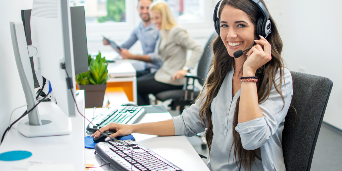 A smiling woman wearing a headset, sitting at a computer in a brightly lit office.