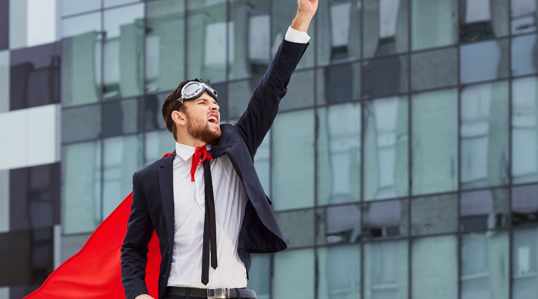 A man in a suit with goggles on his head and a red cape, striking a Superman pose with one arm outstretched, with buildings in the background.