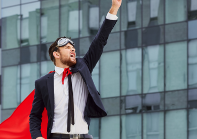 A man in a suit with goggles on his head and a red cape, striking a Superman pose with one arm outstretched, with buildings in the background.