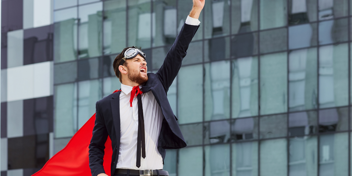 A man in a suit with goggles on his head and a red cape, striking a Superman pose with one arm outstretched, with buildings in the background.