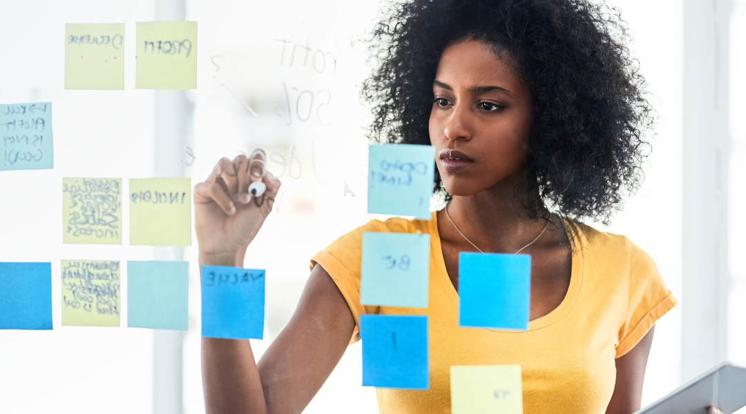 A woman wearing a yellow shirt and writing on a clear backboard that also has blue and yellow sticky notes on it.