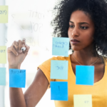 A woman wearing a yellow shirt and writing on a clear backboard that also has blue and yellow sticky notes on it.