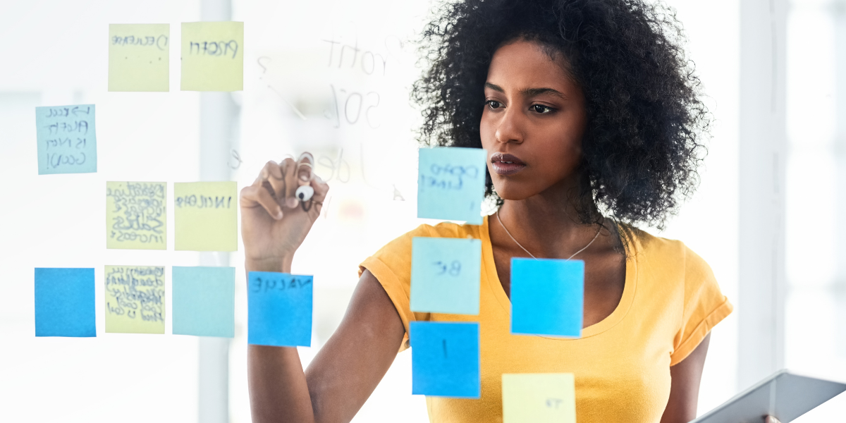 A woman wearing a yellow shirt and writing on a clear backboard that also has blue and yellow sticky notes on it.