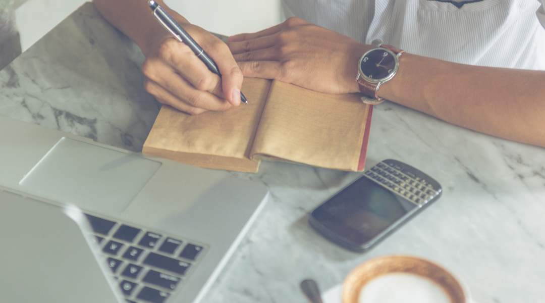 A person at a marble desktop writing in a brown notebook.
