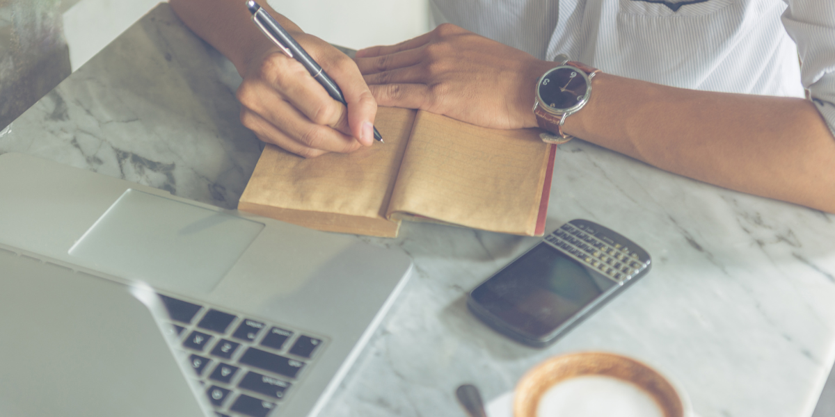 A person at a marble desktop writing in a brown notebook.