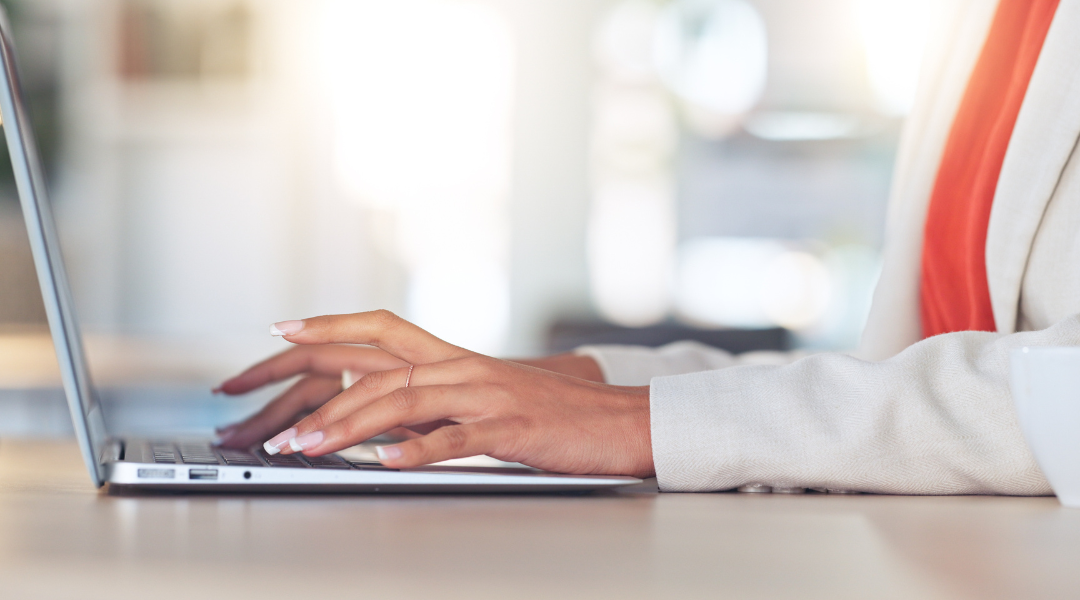 woman wearing a white blazer and orange top working at a laptop.